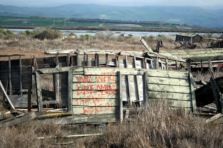 Wreckage of a shack. Graffiti on the shack reads "We were dreaming, then we woke up."