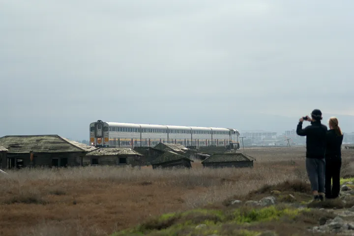 A couple observe a freight train passing Drawbridge, CA as it passes a set of shacks.