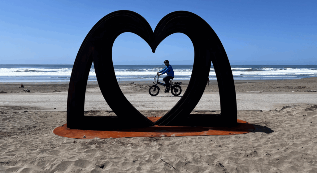 In the foreground, a heart sculpture. Behind it, a bicycle rides past. Behind that, the beach and ocean.