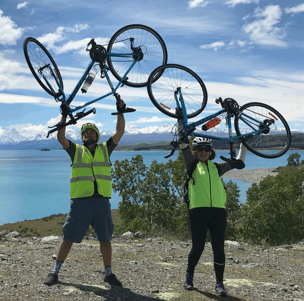 Two bicyclists hold up their bikes in front of a lake in New Zealand.