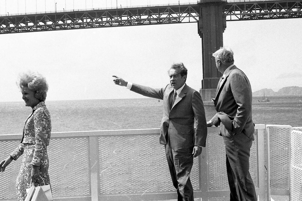 President Richard Nixon stands onboard a boat, pointing to camera left, in front of the Golden Gate Bridge.