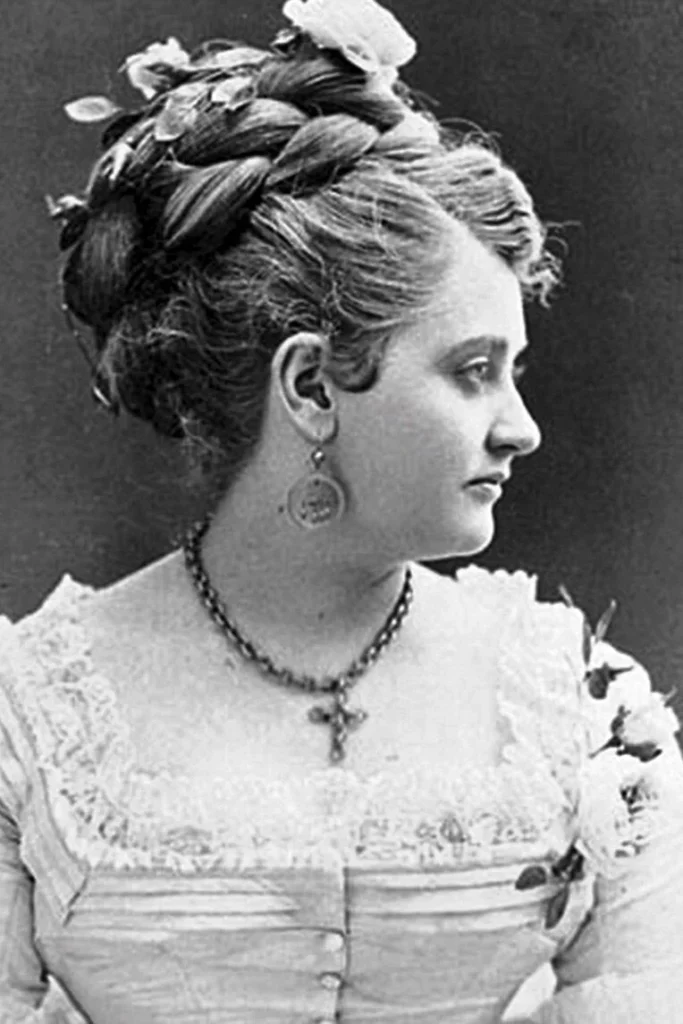 An attractive woman in her 20s dressed in a fine gown. Her hair is done up in a bun. She wears a cross on a chain around her neck.