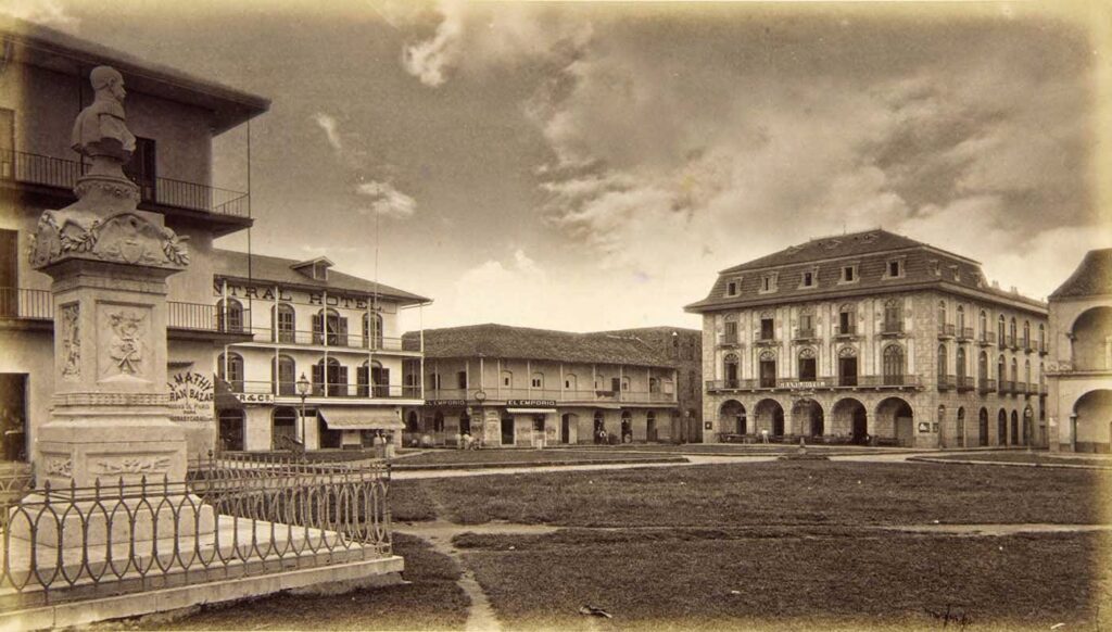 A large plaza with a statue towards the left and several large, grand buildings.