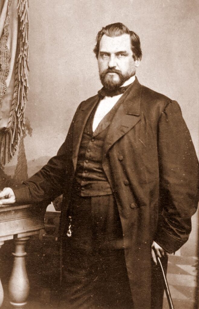 Leland Stanford, a heavy-set, bearded man. He's dressed finely and holds a wooden cane in his left hand.