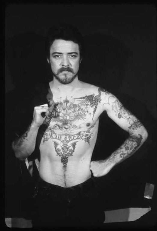 A bearded man stands against a black background. He is shirtless and has elaborate tattoos across his chest and arms.