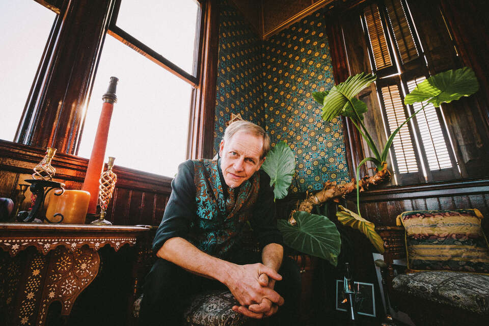 A man sitting in an ornate, stylized, Victorian room leans into camera. He wears a black shirt and a green vest.