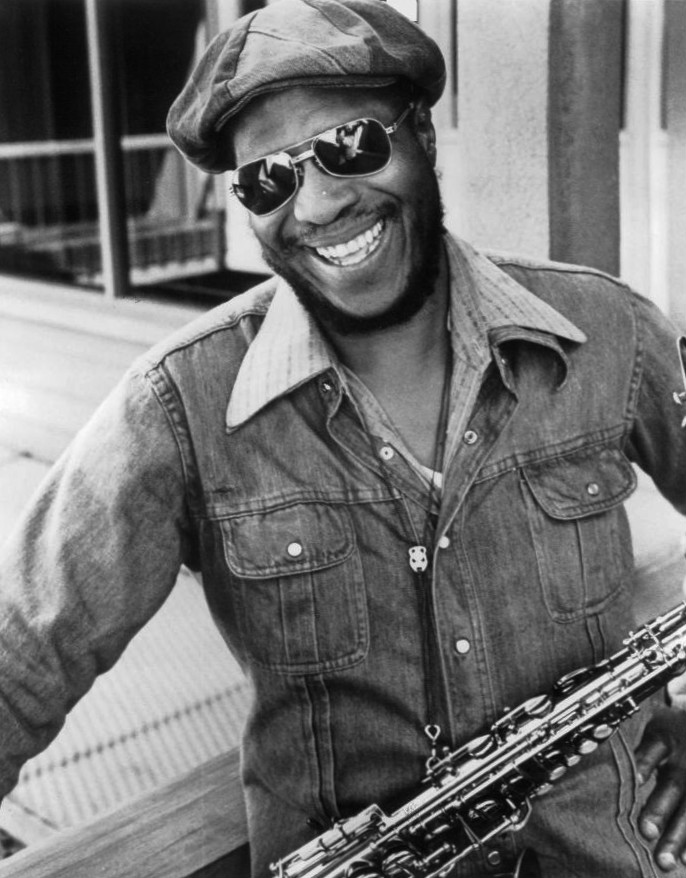 A photo of a smiling man, dressed in denim, wearing sunglasses and a cap. A saxophone is hung over his neck.
